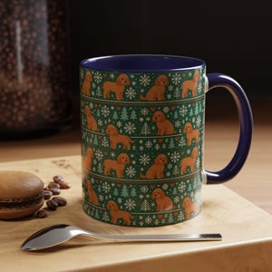May include: A ceramic mug with a dark blue interior and handle. The exterior features a repeating pattern of brown dogs, snowflakes, Christmas trees, and hearts on a green background. A spoon and a pastry are in the foreground.