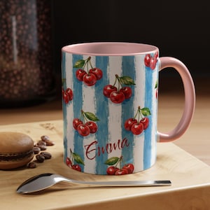 May include: A ceramic mug with a pink handle and interior. The exterior features a blue and white striped pattern with watercolor cherries. The name "Emma" is written in cursive on the mug.