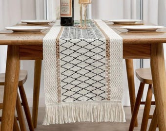 May include: A woven table runner with a geometric diamond pattern in black and white, accented with tan borders and fringe. The runner is displayed on a wooden table set for a meal, with plates, wine, and candles.