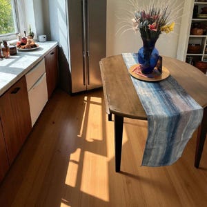 May include: A kitchen scene featuring a wooden table with a blue and white runner. A blue vase with colorful flowers sits on a gold-colored plate. The room has wooden cabinets, a stainless steel refrigerator, and a window.