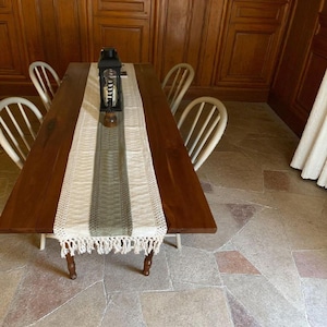 May include: A long, dark wooden dining table with four white chairs. A woven table runner with a central green stripe and fringe detail runs down the center. A black lantern sits on the runner. The floor is made of stone tiles.