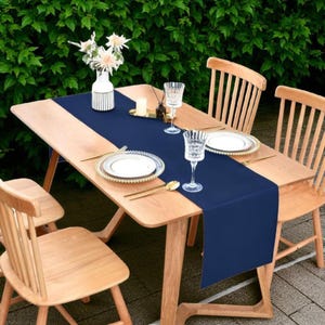 May include: A wooden dining table set for a meal, featuring a navy blue table runner. The table is set with white plates, gold-colored cutlery, and clear wine glasses. Wooden chairs surround the table, with a vase of flowers in the background.