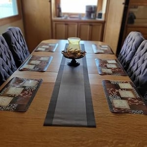 May include: A wooden dining table set with six chairs, placemats, and a centerpiece. The table has a dark gray runner and is set with six placemats with a floral design. A decorative glass candle holder sits on a black stand.