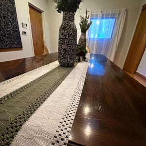 May include: A long, rectangular table runner with a woven design in shades of white and olive green. The runner is placed on a dark wooden table, with decorative vases and greenery in the background. The runner's design includes openwork and textured patterns.