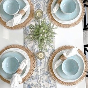 May include: A table setting with four place settings. Each setting includes a light blue bowl and plate, a white napkin, and a woven napkin ring. The placemats are round with a woven border. A floral runner and candles complete the decor.