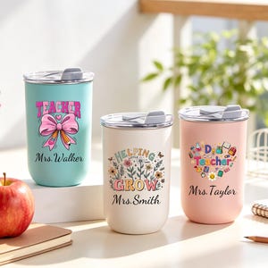 May include: Three stainless steel tumblers with lids, each featuring a unique design. One is teal with a pink bow and the word "Teacher". Another is white with floral graphics and the words "Helping Little Grow". The last is pink with a heart-shaped design and the words "Dear Teacher".