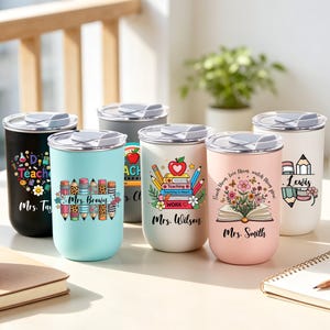 May include: A collection of insulated tumblers with various designs, including pencils, apples, and floral arrangements. Each tumbler features a teacher's name, such as "Mrs. Taylor" or "Mrs. Smith." The tumblers are in black, light blue, white, pink, and gray.