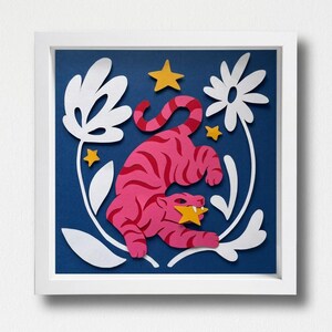 Könnte beinhalten: Ein quadratisches, gerahmtes Papierkunstwerk mit einem rosa Tiger mit dunkelrosa Streifen, der einen gelben Stern im Maul hält. Der Tiger ist von weißen, ausgeschnittenen Blumenformen und zwei weiteren gelben Sternen auf dunkelblauem Grund umgeben.