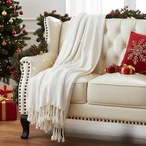 May include: A cream-colored throw blanket with fringe detail draped over a cream-colored tufted sofa. A red pillow with a gold snowflake design and small wrapped gifts are on the sofa. A Christmas tree and garland are in the background.