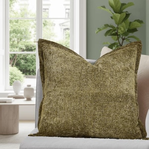 May include: A square, olive-green decorative pillow with a textured weave pattern. The pillow has a darker border and is placed on a light-colored sofa. The background includes a window, a side table, and a houseplant.