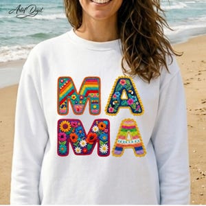 Può includere: Felpa bianca con la scritta "MAMA" in lettere colorate all'uncinetto. Le lettere sono decorate con motivi floreali e arcobaleno. La felpa è uno stile casual e confortevole.