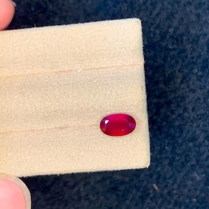 Könnte beinhalten: Ein oval geschliffener Rubin, tiefrot, auf einer beigen Oberfläche präsentiert. Der Edelstein ist vor einem dunkelblauen Hintergrund platziert. Der Rubin wird von den Fingern einer Person gehalten.