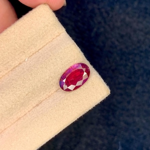 Könnte beinhalten: Ein oval geschliffener, facettierter Rubin-Edelstein mit tiefroter Farbe, der auf einem beigen Stoff liegt. Der Edelstein zeigt innere Facetten und eine polierte Oberfläche, die das Licht reflektiert. Der Hintergrund ist dunkelblau.
