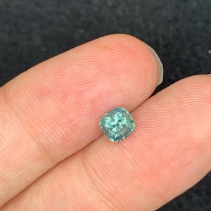 May include: A loose, cushion-cut gemstone with a vibrant teal-green hue and a faceted surface that reflects light. The stone features a square shape with rounded corners and displays internal clarity with visible light-catching facets throughout.