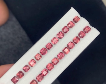 May include: Rows of small, square-cut, pink gemstones are displayed on a white surface. The gems have a vibrant, reddish-pink hue and are arranged in neat lines, showcasing their polished facets. The background is a dark, neutral color.
