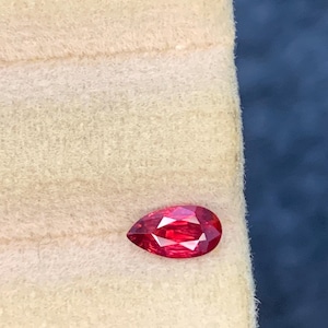 Puede incluir: Una piedra preciosa facetada en forma de pera con un color rojo intenso y vibrante. La piedra presenta un extremo puntiagudo y otro redondeado, con múltiples facetas triangulares y en forma de cometa que reflejan la luz con gran brillo.