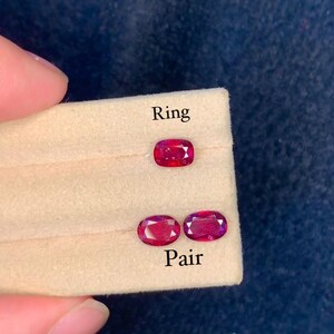 May include: A display of three oval-cut, deep red gemstones. One gemstone is labeled "Ring," and two are labeled "Pair." The gems are set on a beige surface against a dark blue background.