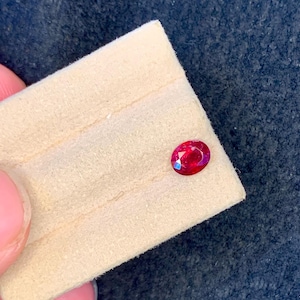 Könnte beinhalten: Ein kleiner, oval geformter, facettierter Rubin-Edelstein. Der Rubin hat eine tiefrote Farbe und liegt auf einem beigefarbenen Hintergrund. Der Edelstein wird von den Fingern einer Person gehalten.