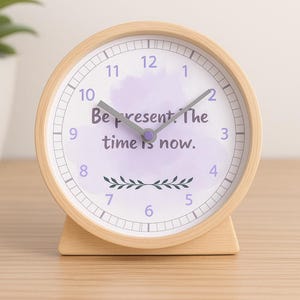 May include: A round wooden alarm clock with a white face and black numerals. The clock face features the text "Be present. The time is now." in a purple watercolor design. The clock is on a wooden surface.