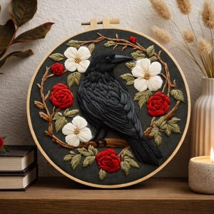 Puede incluir: Bordado de un cuervo negro posado en una rama, rodeado de rosas rojas y flores blancas. El diseño está en un bastidor de madera. El cuervo está sobre una rama, con un fondo gris oscuro. Se exhibe en una estantería de madera.