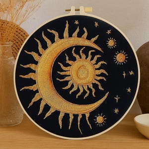 May include: Embroidered celestial wall art featuring a crescent moon and sun design in gold thread on a black background. The embroidery is set in a round wooden hoop, creating a decorative piece for home decor.