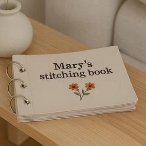 Può includere: Un libro di cucito color crema con il testo "Mary's stitching book" e due illustrazioni di fiori arancioni. Il libro è rilegato con anelli argentati e poggia su una superficie di legno chiaro. Un piccolo vaso bianco con fiori secchi è sullo sfondo.