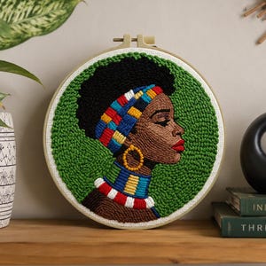 May include: A circular embroidery hoop art piece featuring a punch needle portrait of a person with dark skin and black hair, wearing a colorful patterned headband, a gold hoop earring, and a multi-colored neck collar against a green background.
