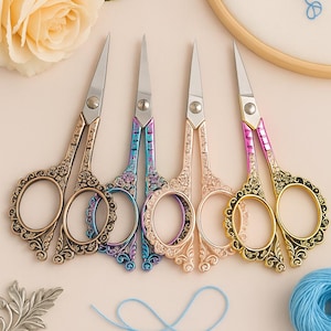 Pode incluir: Cinco tesouras decorativas de bordado com cabos ornamentados em ouro, bronze, rosa e azul. As tesouras têm lâminas prateadas afiadas e estão dispostas sobre uma superfície clara. Uma bola de lã azul e uma flor também são visíveis.