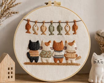 Può includere: Ricamo su telaio raffigurante cinque gatti con maglioni sotto una fila di pesci ricamati. I gatti sono neri, bianchi e arancioni, con vari disegni di maglioni. Il telaio è posto su una mensola di legno con una statuina di gatto in ceramica.