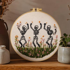 May include: An embroidered hoop art piece showing three dancing skeletons. The skeletons are black and white, set against a cream fabric. They are surrounded by embroidered flowers in pink, orange, and green. The wooden hoop holds the artwork, which is a circular design.