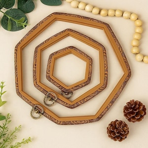 May include: Three wooden embroidery hoops in varying sizes, arranged in a nested pattern. The hoops are a warm brown with a wood grain design. Antique brass hardware is attached to the largest hoop. Two small pinecones and a string of wooden beads complete the arrangement.