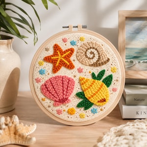 Könnte beinhalten: Ein runder Stickrahmen aus Holz mit einem cremefarbenen Stoffeinsatz, der texturierte Punch-Needle-Motive eines orangefarbenen Seesterns, einer beigen Spiralschnecke, einer rosa Muschel und einer gelben Muschel mit grünen Blättern sowie kleinen Blüten zeigt.