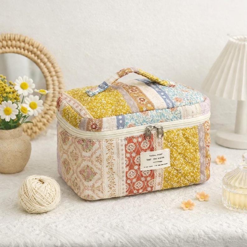 May include: A patchwork cosmetic bag with a handle, featuring a variety of floral and geometric patterns in yellow, blue, and red. The bag has a white zipper and a small tag with text. A ball of twine and flowers are in the background.