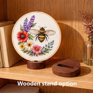 May include: A round embroidery hoop featuring a detailed bee design surrounded by colorful flowers and foliage. The embroidery is mounted on a wooden stand. The text "Wooden stand option" is visible at the bottom.