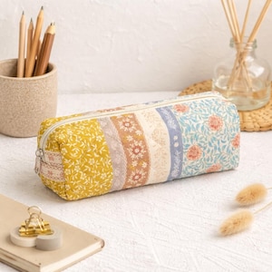 May include: A rectangular pencil case with a zipper closure. The case is made of quilted fabric with a patchwork design featuring floral and patterned panels in yellow, lavender, cream, and blue. A small gold paper clip and a notepad are in the foreground.