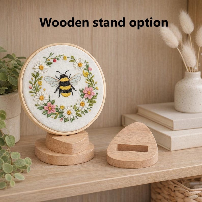 May include: A round, embroidered artwork featuring a bee surrounded by flowers, displayed on a wooden stand. The text "Wooden stand option" is above the artwork. The stand is made of light-colored wood.