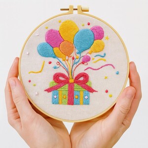 May include: Embroidered hoop art featuring a colorful birthday design. The artwork showcases a gift box with a red bow and a cluster of balloons in shades of yellow, pink, and blue. The design is set against a neutral fabric background.