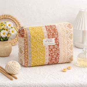 May include: A rectangular cosmetic bag with a patchwork design. The bag features vertical stripes in yellow, rust, and cream colors with floral and geometric patterns. A small label reads "today, mood feel the warm."
