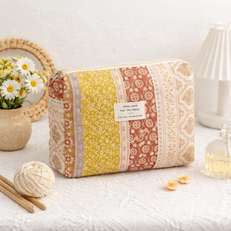 May include: A rectangular cosmetic bag with a patchwork design. The bag features vertical stripes in yellow, rust, and cream colors with floral and geometric patterns. A small label reads "today, mood feel the warm."