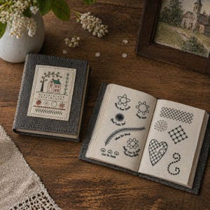 Puede incluir: Un libro cubierto de tela gris con un diseño cosido de una casa y un árbol. El libro abierto muestra varios patrones de puntadas de bordado en hilo negro. Los libros están sobre una superficie de madera, con un jarrón de flores blancas en el fondo.