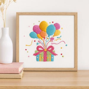 May include: Embroidered artwork in a wooden frame. The image features a gift box tied with a pink bow, with colorful balloons rising above it. The balloons are yellow, blue, pink, and orange. Confetti and streamers add to the festive design.