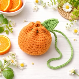 Könnte beinhalten: Ein gehäkelter, orangenförmiger Beutel mit Kordelzug, grünem Blatt und Stiel. Die Tasche ist leuchtend orange, Blatt und Stiel sind grün. Orangenstücke und Gänseblümchen sind im Hintergrund.