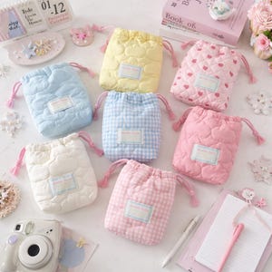 Peut inclure: Collection de pochettes à cordon de serrage aux couleurs pastel. Les pochettes sont matelassées avec des motifs de cœurs ou de carreaux. Les couleurs incluent le bleu clair, le jaune, le rose et le blanc. Chaque pochette a une petite étiquette et un cordon de serrage rose.