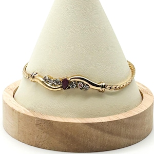 May include: A gold-toned bracelet featuring a twisted chain band and a central curved decorative element. The center piece is set with a single marquise-cut red stone flanked by two clear round stones on each side, all set in a polished gold metal.