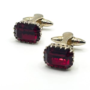 May include: A pair of rectangular cufflinks featuring deep red, faceted glass stones set in silver-toned metal frames. Each cufflink has a prong setting around the stone and a hinged toggle mechanism on the back for securing through a shirt cuff.