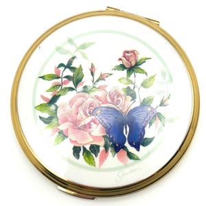 May include: A round, gold-toned compact mirror with a floral and butterfly design. The center features pink roses and a blue butterfly against a white background, framed by a gold rim. The compact is closed.