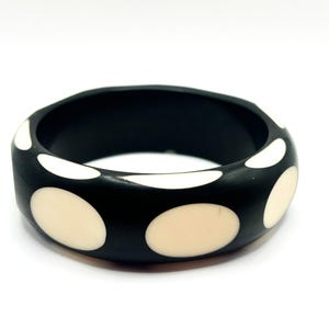 May include: A thick, circular bangle bracelet featuring a matte black base with inlaid, smooth, cream-colored oval shapes spaced evenly around the exterior surface. The bracelet has a wide, solid band construction with a polished finish.
