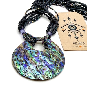 Puede incluir: Un collar llamativo con un gran colgante de concha de abulón iridiscente. El colgante está unido a múltiples hilos de cuentas negras. Una etiqueta con un diseño de ojo y el texto "BD-EYE Boutique" es visible.