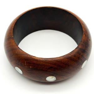 May include: A wide, circular bangle bracelet crafted from dark brown wood with a smooth, polished finish. The exterior surface of the bracelet is inlaid with several small, round, white mother-of-pearl accents spaced evenly around the circumference.