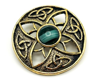Puede incluir: Broche redondo dorado con una piedra verde central. El broche presenta un diseño calado con patrones de nudos celtas y un borde texturizado. La piedra central es de color verde oscuro con remolinos verdes más claros. Sobre fondo blanco.